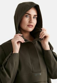 Vrouw met blauwe ogen die een donkergroene capuchonjas draagt en de hoedranden dicht bij haar gezicht houdt tegen een effen witte achtergrond.