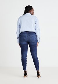 Egy fonott hajú nő kék csíkos pólót, sötétkék skinny farmert és fekete magassarkú cipőt visel, háttal állva.