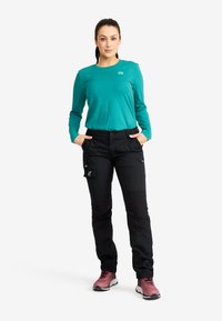 Kvinna i teal långärmad tröja, svarta cargobyxor och rosa sneakers, står med händerna i fickorna mot vit bakgrund.