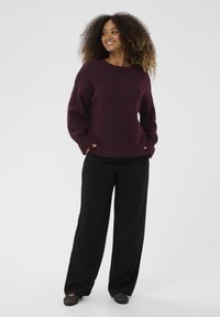 Burgundiško spalvos per didelis megztinis iš minkštos medžiagos, derinamas su juodomis plačioms kojoms kelnėmis ir juodais tekstūruotais bateliais. Modelis stovi su rankomis kišenėse.