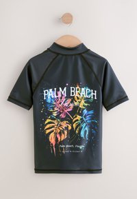 Juodas trumpomis rankovėmis rash guard'as su spalvingais palmės lapų raštais, atrodantys kaip išsilieję dažai. Apačioje parašyta „Palm Beach, Florida“.