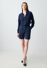 Marineblaue Leinenbluse mit zwei Brusttaschen, geknöpft vorne, kombiniert mit passenden taillierten Shorts. Das Model trägt dunkelbraune Absatzschuhe.