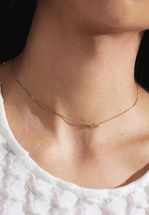Gul kædedyse med justerbar længde, prydet med ordet "Søster" i en flydende skrift. Simpelt og delikat design mod en tekstureret hvid stof.