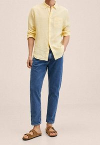 Chemise jaune en lin à boutons, jean bleu et sandales en daim marron. La chemise possède une poche et des manches retroussées, le jean est légèrement fuselé.