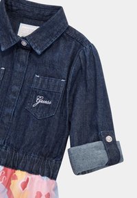 Sukienka dżinsowa z ciemnoniebieską górą, dwiema kieszeniami na piersiach i podwiniętymi rękawami. Spódnica ma kolorowy wzór w odcieniach różowego i pomarańczowego.