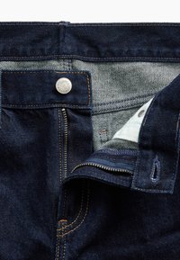 Σκούρα μπλε τζιν από denim με μερικώς ανοιχτό φερμουάρ, ορατό κουμπί και αντίθετη πορτοκαλί ραφή στη μέση και την περιοχή του φερμουάρ.