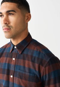 Brązowo-niebieska koszula flanelowa w kratę z kołnierzykiem na guziki, pojedynczą kieszenią na piersi oraz białymi guzikami na teksturowanej tkaninie.