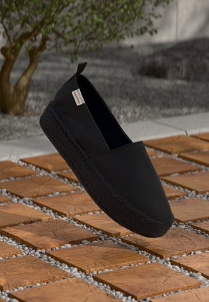 Czarne buty wsuwane z grubą, teksturowaną podeszwą unoszące się nad drewnianymi kwadratowymi kafelkami z przerwami wypełnionymi żwirem, małe drzewo na rozmytym tle.