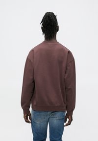 Ognjen rjav pulover z mehko teksturo in preveliko prileganje, z rebrastim izrezom in manšetami, kombiniran s modrimi denim kavbojkami.