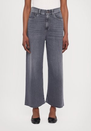 Vrouw draagt hoge taille grijze wijde jeans met onafgewerkte zoom, gecombineerd met zwarte puntige platte schoenen, staand tegen een effen achtergrond.