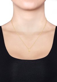 Goldene Halskette mit einer dünnen Kette, die einen kleinen Anhänger in Form des Blockbuchstabens "L" trägt. Die Kette wird über einem schwarzen Oberteil getragen und vor einem schlichten Hintergrund präsentiert.