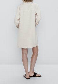 Beiges Leinenhemdkleid mit knielangem Schnitt, geknöpfter Rückseite und langen, umgeschlagenen Ärmeln. Kombiniert mit schwarzen flachen Sandalen.