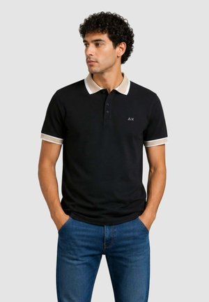 Homme portant un polo noir avec col et bords de manches beige, debout les mains dans les poches, regardant vers la gauche.