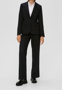 Czarny dopasowany garnitur z pojedynczym guzikiem w marynarce, prostymi spodniami i błyszczącymi czarnymi butami. Teksturowana tkanina z kieszeniami.