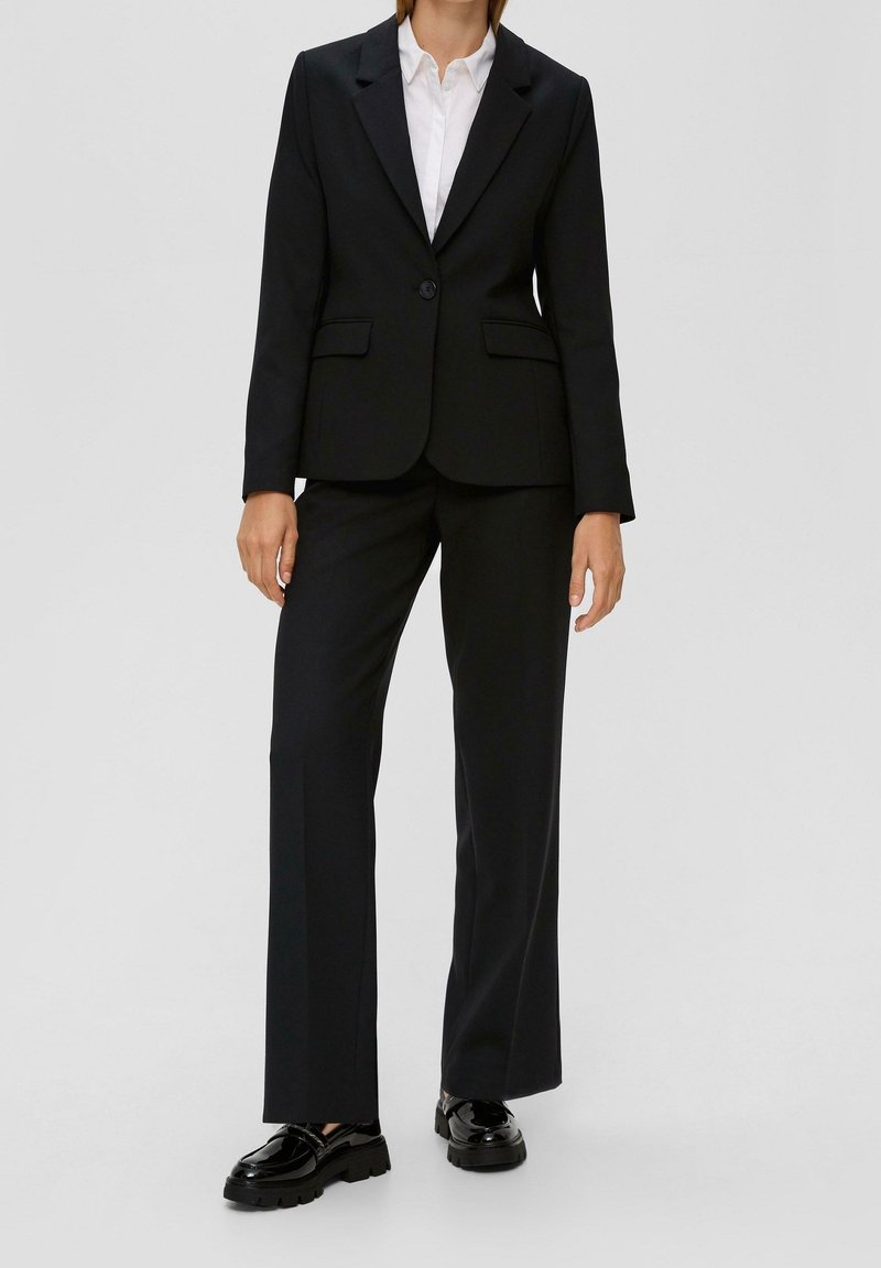Czarny dopasowany garnitur z pojedynczym guzikiem w marynarce, prostymi spodniami i błyszczącymi czarnymi butami. Teksturowana tkanina z kieszeniami.