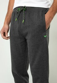 Graue Jogginghose aus einem weichen Stoff mit einem dehnbaren Bund und grünen Akzenten, zwei Seitentaschen und einem Logo am Oberschenkel.