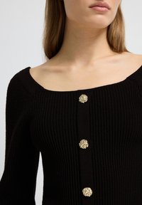 Schwarzes, geripptes Oberteil mit weit ausgeschnittenem Hals, langen Ärmeln und drei kunstvoll gestalteten goldenen Knöpfen auf der Vorderseite, das einen strukturierten Stoff hervorhebt.