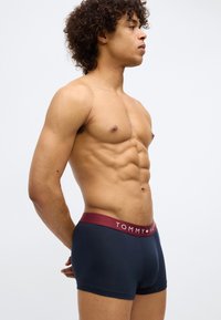 Muškarac bez košulje s kovrčavom kosom, obučen u mornarske Tommy Hilfiger bokserice s bordo pojasom, stoji u profilu na jednobojnoj pozadini.