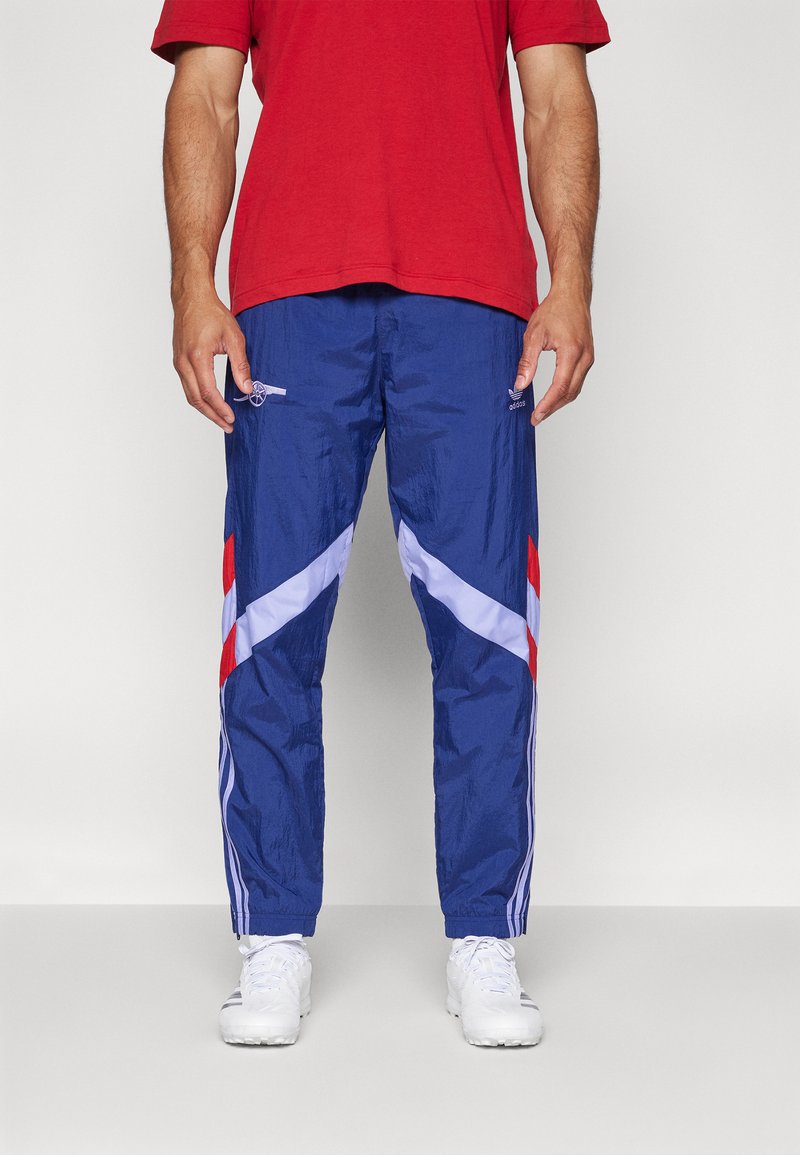 Modré lehké sportovní kalhoty s červenými a bílými diagonálními pruhy, elastickým pasem a síťovinou. Nosené s červeným tričkem a bílými botami.