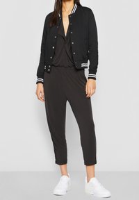 Černá bomber bunda s bílými pruhy, nošená přes tmavý overal. Bílá sneakers doplňují celý outfit. Hladká tkanina s uvolněným střihem.