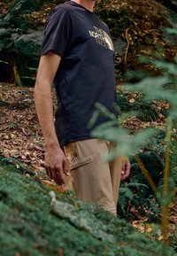 Black short-sleeve t-shirt with "The North Face" logo and khaki cargo pants with a side pocket, set against a forest background.