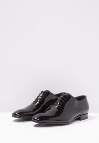 Chaussures en cuir verni noir au design épuré, à bout rond et à fermeture à lacets. Texture lisse et détail de talon minimaliste.