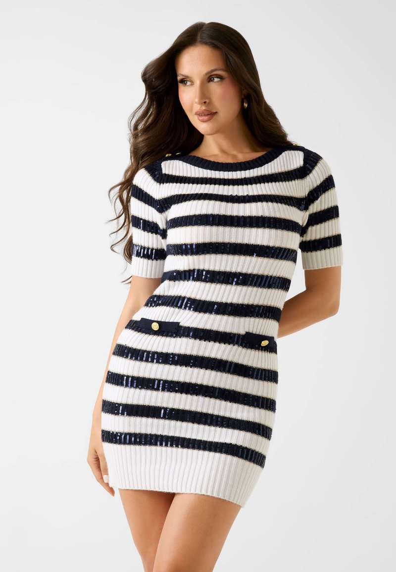 Kobieta nosząca krótką, prążkowaną, dzianinową mini sukienkę z poziomymi czarno-białymi paskami i złotymi guzikami na przednich kieszeniach.