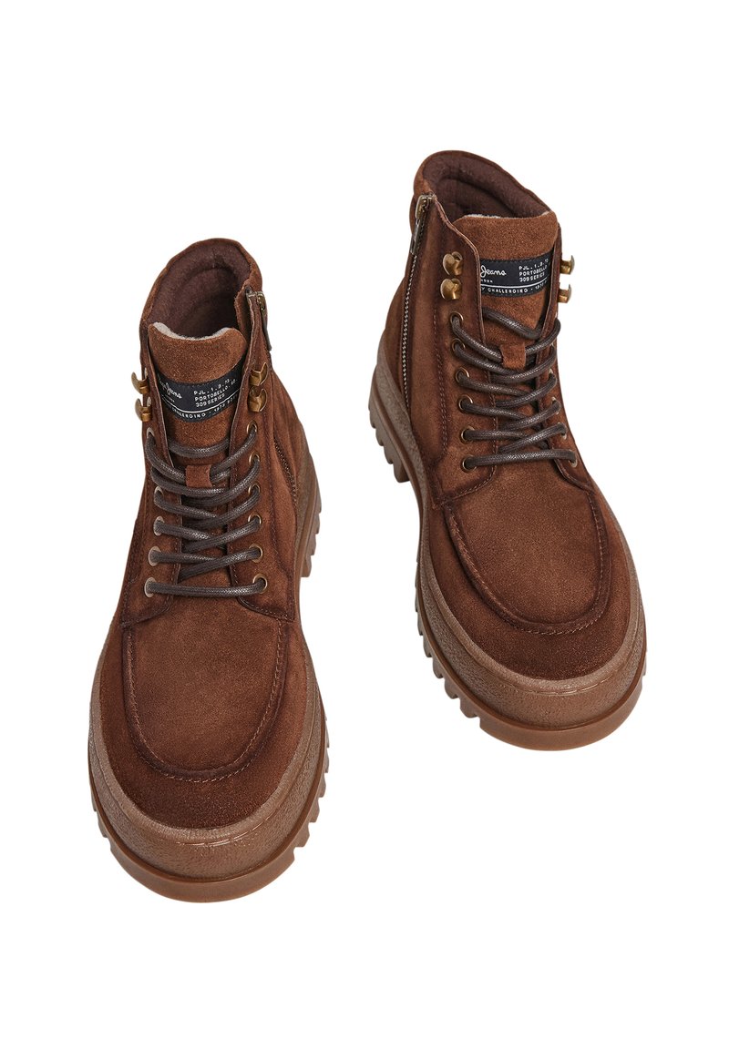 Botas de tobillo de ante marrón con herrajes de metal, cierre con cordones, detalle de cremallera, cuello acolchado y suelas de goma texturizadas.