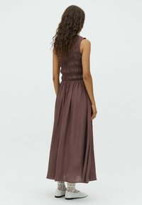 Maxiklänning i djup mauve, med smockad byst, flödande kjol och textil med struktur. Matchas med vita skor och spetsstrumpor.