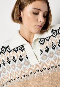 Sweter dzianinowy z białym kołnierzykiem. Posiada geometryczny wzór w kolorze czarnym, brzoskwiniowym i kremowym. Miękka tekstura i luźny krój.