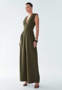 Olivově zelený overal s hlubokým výstřihem do V, krátkými rukávy a širokými nohavicemi. Látka vypadá hladce s viditelnými švy. Černé sandály na podpatku.