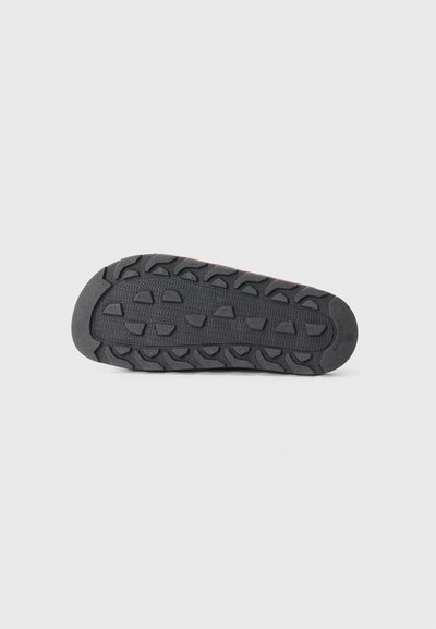 Semelle de chaussure en caoutchouc avec une surface noire texturée, présentant un motif de grip prononcé pour une meilleure adhérence et stabilité. Une petite ligne d'accent rouge est visible.
