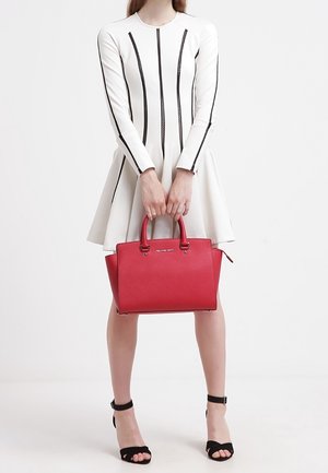 Frau in weißem Langarmkleid mit schwarzen Streifen hält eine rote Handtasche und trägt schwarze Absatzschuhe mit Knöchelriemen vor einem schlichten weißen Hintergrund.
