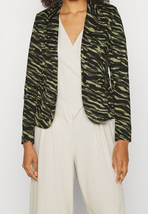 Žena v zeleno-černém blejzru s tygřím vzorem přes béžový overal s výstřihem do V, stojící s rukama volně podél těla.