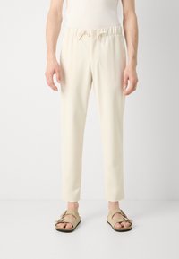 Pantalones elásticos de color beige claro con cintura ajustable, piernas estrechas y textura de tela suave, combinados con sandalias color marrón claro con doble hebilla.