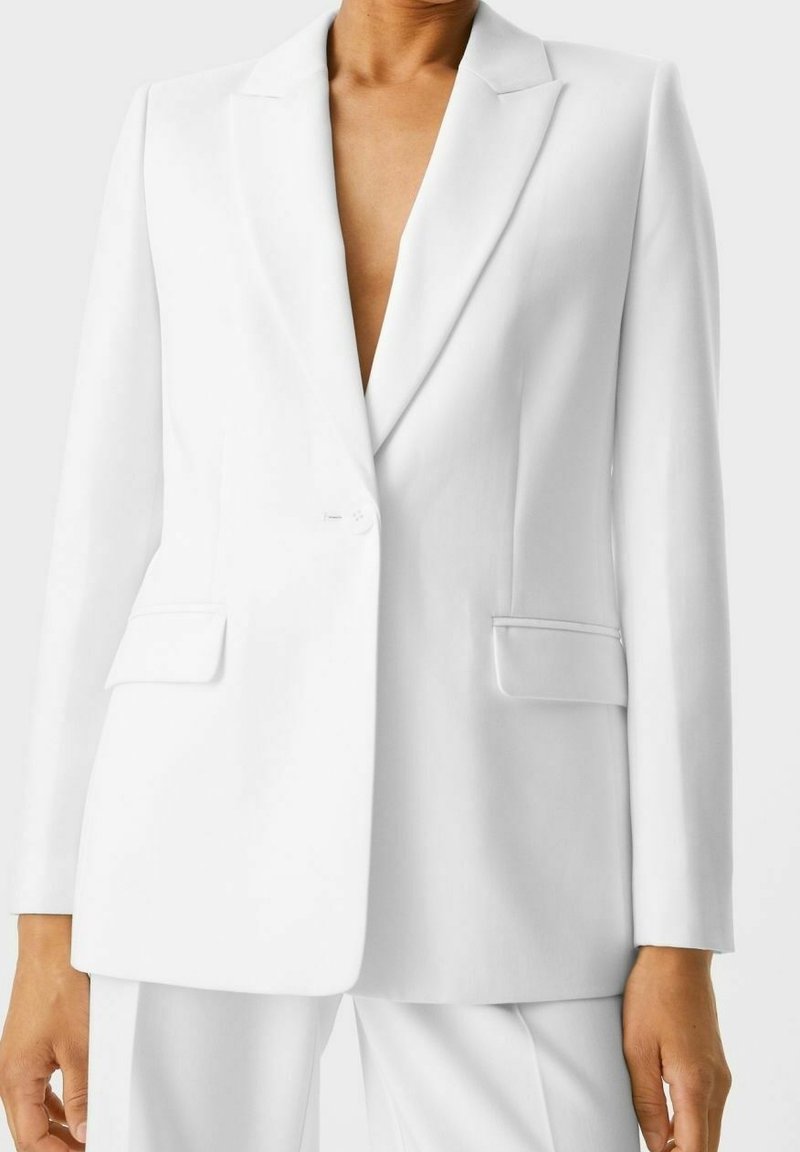 Osoba ubrana w dopasowaną białą marynarkę z jednym guzikiem, z klapami, kieszeniami z patkami i dopasowanymi białymi spodniami.