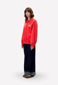 Sweatshirt rouge avec un col polo, trois boutons et un patch logo. Associé à un jean large en denim foncé et des chaussures slip-on imprimé léopard.