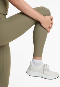 Leggings ribbați de culoare verde măsliniu purtați cu încălțăminte sport albă, având un material texturat și talpă căptușită, asortați cu o șosetă albă până la gleznă.
