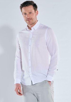 Man with short brown hair wearing a long-sleeve white button-up shirt and light gray pants, standing with one hand in pocket.