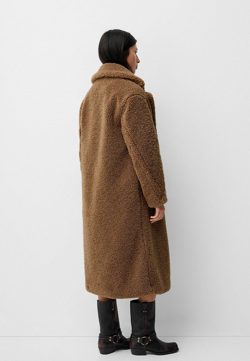 Femme aux cheveux foncés portant un long manteau en teddy marron et des bottes noires jusqu'aux mi-mollets, debout et faisant face à un mur blanc.