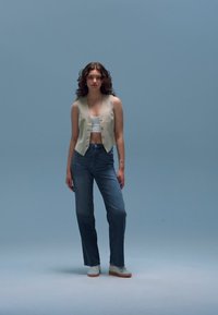 Šviesiai pilkas berankovis liemenė virš baltos apkarpyto stiliaus palaidinės, derinamas su aukšto liemens aukštaūgiais mėlynais džinsais ir baltais sportiniais bateliais. Lygios tekstūros, laisvas kasdienis stilius.