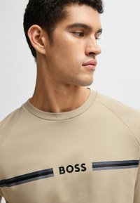 Beige sweatshirt with a crew neck and raglan sleeves, featuring black and gray horizontal stripes and "BOSS" logo in the center.