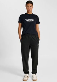 Marineblå t-shirt med korte ærmer og hvid "hummel"-logo, kombineret med sorte joggers med sidelommer og elastisk taljebånd. Beige sneakers.