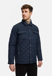 Giubbotto trapuntato blu navy con motivo a diamante, bottoni a pressione, due tasche sul petto e colletto. Tessuto liscio con una texture morbida.
