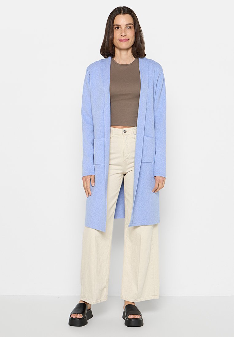 Frau steht in einem hellblauen, knielangen Cardigan, braunem Crop-Top, cremefarbenen, weit geschnittenen Hosen und schwarzen Plateausandalen vor einem weißen Hintergrund.