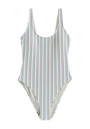 Young woman with long brown hair wearing a sleeveless white top with thin vertical stripes, standing outdoors in sunlight.