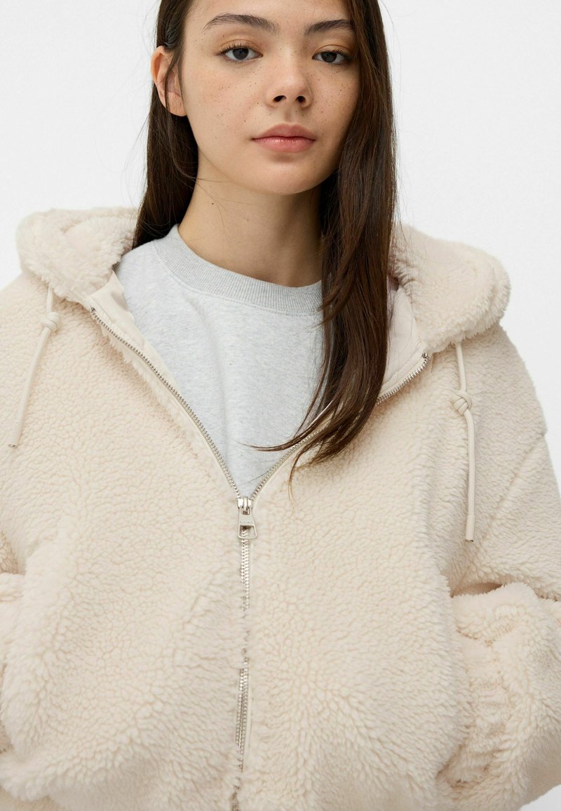 Sweat à capuche en polaire couleur crème avec une surface douce et texturée. Comprend une fermeture éclair, une capuche avec cordon de serrage et des poches avant. Porté par-dessus une chemise grise.