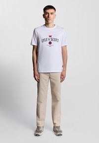 T-shirt bianco in cotone con logo blu "LYLE & SCOTT" e grafica di una rosa rossa; abbinato a pantaloni beige chiaro e sneakers grigie.