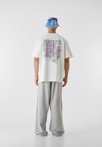 Biela voľná džersejová tričko s grafickým dizajnom a nápisom "STRATENÁ MLADOSŤ" v purpurovej farbe na chrbte, skombinované so svetlošedými teplákmi a viacfarebným klobúkom.