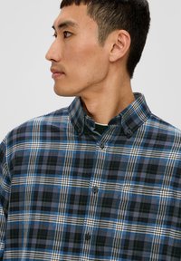 Chemise à boutons en tissu coton avec un motif en plaid bleu, noir et blanc. Col standard avec fermeture à boutons et poche poitrine.